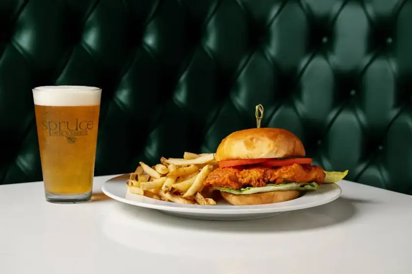 A crispy hot chicken sandwich with fries beside a Spruce on Blackbridge pint of beer